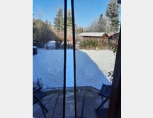 Blick durch eine geöffnete Fliegengittertür auf einen schneebedeckten Garten mit Bäumen, Sträuchern und einer kleinen Holzhütte im Hintergrund. Rechts und links vor der Tür stehen schwarze Klappstühle, und der Himmel ist klar und blau.