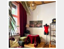 Wohnzimmer mit dekorativer Einrichtung: Ein Raum mit roten Vorhängen, einem runden Tisch mit roter Tischdecke und einem Sitzbereich neben einem großen Fenster. Im Hintergrund eine Schreibmaschine, ein Wandbild mit Flamingos, ein glitzernder Kronleuchter an der Decke und eine Schaufensterpuppe mit rotem Badeanzug.