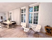 Heller Wohnraum mit großen, weiß gerahmten Fenstern, die teils von Vorhängen bedeckt sind. Der Raum verfügt über Holzboden, einen Esstisch mit Stühlen, moderne Sessel und ein kleines Glas-Beistelltischchen, mit Blick ins Grüne draußen.