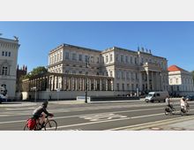 Historisches Gebäude im klassizistischen Stil mit Säulengang und reich verzierten Fassaden, flankiert von weiteren Gebäuden. Im Vordergrund verlaufen eine Straße mit Fahrradfahrern und ein Fußweg, bei sonnigem Wetter.