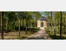 Tree-lined gravel path in a park leading to a small octagonal pavilion with pale orange walls, arched openings, and a dark domed roof. The pavilion sits on a grassy lawn surrounded by tall trees and scattered classical statues.