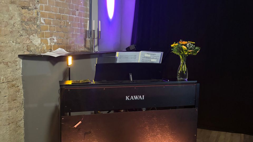 Alt text: A black upright digital piano sits on a wooden floor against an exposed brick wall and dark curtain backdrop, with purple uplighting on the wall. Sheet music is propped on the stand, and a small lamp and a vase of flowers rest on top of the piano.