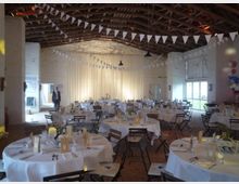 Ein dekorierter Veranstaltungssaal in einer rustikalen Scheune mit Holzbalkendecke und weißen Wänden. Runde Tische mit weißen Tischdecken und gedecktem Geschirr stehen im Raum, während Wimpelgirlanden quer durch den Saal hängen; im Hintergrund hängt ein halbtransparenter Vorhang, der den Raum teilt.