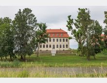 Historisches Gebäude mit rotem Satteldach und cremefarbener Fassade, umgeben von Bäumen und einer weitläufigen Grünfläche, im Vordergrund ein Fluss oder Teich. Eine steinerne Treppe führt zum erhöht gelegenen Eingang des Gebäudes.