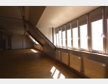 Großer, leerer Dachraum mit schrägen Decken und einer Fensterreihe auf der rechten Seite, durch die Tageslicht einfällt. Der Boden besteht aus hellbraunem Holz, und Heizkörper sind unter den Fenstern entlang der Wand angebracht.