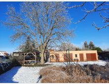 Hof mit schneebedecktem Boden, einer großen, kahlen Baumkrone im Vordergrund und einem beigefarbenen Gebäude mit mehreren Holzgaragentoren im Hintergrund. Rechts steht ein kleiner Carport, links ein offener, überdachter Holzunterstand.