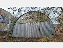 Blick auf eine halbrunde Lager- oder Industriehalle mit gewölbtem Dach aus Wellblech und einer breiten Front aus senkrechten Metallpaneelen. Im Vordergrund liegen braches, teils überwuchertes Gelände und kahle Baumäste; links grenzt ein älteres Gebäude mit großen Fenstern an.