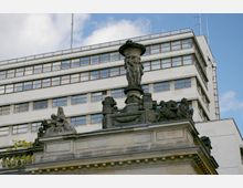 Historische Steinbalustrade mit detailreichen Skulpturen, darunter Figuren und ein zentral platzierter Putto mit einer Vase, vor einem modernen Gebäude mit weißer Fassade und großen Fenstern. Der Himmel ist leicht bewölkt.