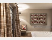 Zimmer mit heller, beiger Wandfarbe, karierten Vorhängen und Parkettboden. An der Wand hängt ein Kunstwerk mit Campbell's-Tomatensuppendosen, darunter ein Stuhl mit einem bunten Kissen.