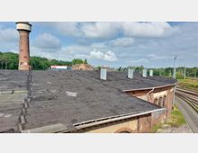 Industriegebäude mit flachem Dach und mehreren Lüftungsschächten, im Hintergrund ein roter Backstein-Wasserturm und eine Bahnanlage mit Gleisen, umgeben von Grünflächen und vereinzelten Gebäuden.