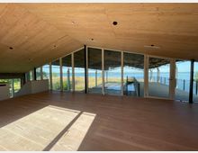 Innenraum eines Gebäudes mit hellem Holzfußboden und einer Holzdecke, mit großen Glasfronten, die einen Blick auf Wasser, Schilf und eine Außenterrasse ermöglichen. Die Umgebung wirkt offen und naturnah mit Sicht auf eine Landschaft am See.