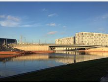 Moderne Bürogebäude mit einer hellen, rechteckigen Fassadenstruktur stehen neben einer Brücke über einen Kanal. Im Hintergrund ist ein gläsernes Gebäude mit gewölbtem Dach zu sehen, das möglicherweise ein Bahnhof ist.