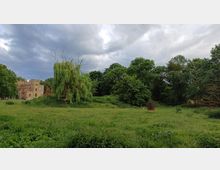 Weite Wiese mit hohem Gras, umgeben von Bäumen, darunter eine große Trauerweide in der Bildmitte. Links steht ein historisches Gebäude aus hellem Stein mit roten Akzenten, im Hintergrund dichter Baumbestand unter einem bewölkten Himmel.