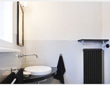 Minimalistisches Badezimmer mit weißen Wänden, einem runden Waschbecken auf einer schwarzen Ablage, einer wandmontierten Armatur, einem schwarzen Handtuchheizkörper und einer kleinen Ablage mit Handtuchhaken.