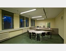 Ein einfacher Konferenzraum mit gelb gestrichenen Wänden und mehreren rechteckigen Tischen, die zu einem unregelmäßigen Muster angeordnet sind. Der Raum verfügt über grüne Kunststofffenster, eine weiße Heizung, eine Wanduhr und an der Decke montierte Leuchtstofflampen.
