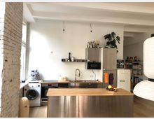 Moderne Küche mit einer Kücheninsel aus Edelstahl und Holzarbeitsplatte, umgeben von weißen Wänden und einem freiliegenden Mauerwerk. Im Hintergrund sind ein Kühlschrank, Schränke, Regale, eine Waschmaschine und eine Fensterfront zu sehen; Dekoration wie eine Topfpflanze, Küchenutensilien und ein Wandkalender ergänzen den Raum.