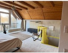 Ein Dachzimmer mit geneigter Holzdecke und großen Fenstern, die viel Tageslicht hereinlassen. Das Zimmer enthält ein Bett, einen Teppich, eine Zimmerpflanze, eine Sitzgelegenheit sowie eine gelb gestrichene Treppe, die nach unten führt.