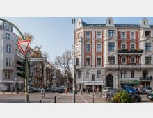 Straßenecke in einer Stadt mit einem historischen, prächtig verzierten Gebäude im Gründerzeitstil mit roten und weißen Fassaden sowie Balkonen. Im Vordergrund Straßenschilder der "Knesebeckstraße" und "Goethestraße", eine Verkehrsampel, parkende Autos, Fahrräder und Fußgänger bei sonnigem Wetter.