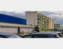 Mehrstöckige Wohngebäude mit bunter Fassade und modernen Fenstern, daneben ein flacher Bau mit blauer und weißer Außenwand. Im Vordergrund ist ein Parkplatz mit parkenden Autos zu sehen, im Hintergrund bewölkter Himmel.