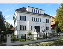 Weiße zweistöckige Stadtvilla mit dunkelgrauem Walmdach und Fensterläden, umgeben von einem gepflegten Garten und einem schwarzen Metallzaun. Das Gebäude verfügt über eine breite Vortreppe mit blauem Blumentopf und ist in einem ruhigen Wohngebiet gelegen.