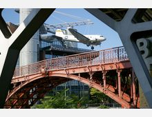 Außenansicht des Deutschen Technikmuseums in Berlin mit einer historischen Douglas C-47-Flugzeugattrappe, die scheinbar über dem Gebäude schwebt, und einer dekorativen roten Stahlbrücke im Vordergrund. Im Hintergrund sind moderne Glas- und Stahlelemente des Museums sichtbar.
