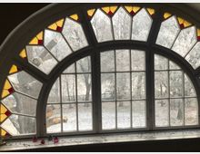 Rundes Fenster mit weißen Rahmen und dekorativen Buntglas-Elementen in Gelb und Rot, blickt auf einen schneebedeckten Garten mit kahlen Bäumen und einem Holzzaun im Hintergrund. Auf der Fensterbank stehen kleine Kerzenhalter und Lichterketten.