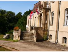 Beige gestrichenes, historisch wirkendes Gebäude mit hohen Fenstern und einem kleinen Balkon über einem Eingangsbereich mit Steinbögen. Eine Treppe führt zum Eingang hinauf, daneben sind rote und weiße Flaggen angebracht, und im Hintergrund ist eine grüne, bewaldete Landschaft sichtbar.