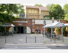 Außenansicht des S-Bahnhofs Berlin-Schlachtensee mit Ziegelfassade, einem Torbogen-Eingang und einem Schild mit dem Stationsnamen. Im Vordergrund befinden sich Fahrradständer mit abgestellten Fahrrädern sowie eine überdachte Abstellfläche und ein Zebrastreifen.