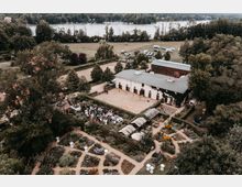Luftaufnahme eines Landguts mit Gartenanlage und weißen Gebäuden, umgeben von Wiesen und Bäumen. Im Vordergrund ein gepflegter Garten mit Blumenbeeten und Wegen, im Hintergrund ein Fluss mit Waldufer.
