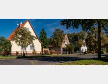 Straßenszene mit freistehenden Einfamilienhäusern in einer Wohngegend, umgeben von Bäumen und kleinen Vorgärten; die Häuser haben spitze Giebel und teilweise rote oder dunkle Ziegeldächer. Im Vordergrund ist eine asphaltierte Straße mit grünem Rasenstreifen sichtbar.