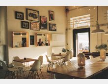 Heller Innenraum eines Cafés mit minimalistischer Einrichtung, bestehend aus Holztischen und weißen Stühlen. An der Wand hängen gerahmte Bilder, darunter drei Fensteröffnungen zum Tresen, wo eine Person steht; unter warmem Licht sind Pflanzen und eine offene Tür sichtbar, die nach draußen führt.