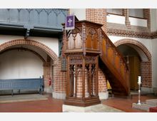 Holzkanzel in einer Kirche mit detaillierten gotischen Schnitzereien, flankiert von gemauerten Rundbögen und Wänden aus roten Ziegelsteinen; daneben eine blaue Holzbank und eine Tafel mit Liednummern.
