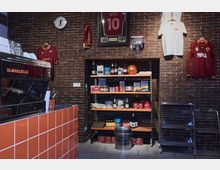 Innenansicht eines Cafés oder einer kleinen Ladenfläche mit Backsteinwand. Es sind Regale mit Lebensmitteln und Getränken, eingerahmte Fußball-Memorabilien sowie ein orange gefliester Tresen im Vordergrund und eine Kaffeemaschine zu sehen.