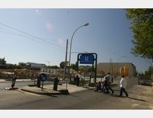 Straßenszene mit einer Baustelle neben der U-Bahn-Station "Schwartzkopffstraße", erkennbar durch ein blaues Stationsschild. Im Hintergrund sind Sandhaufen, Bauzäune, Kräne und Gebäude zu sehen, während Passanten mit Kinderwagen eine Straße überqueren.