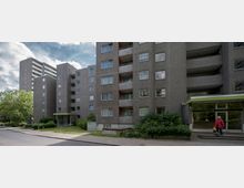 Mehrstöckiges Wohngebäude aus Beton mit Balkonen und zahlreichen Fenstern, umgeben von begrünter Fläche und Gehwegen; eine Person steht vor dem Eingang mit einem Vordach. Im Hintergrund ist ein höherer Wohnkomplex zu sehen.