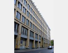 Ein mehrstöckiges Bürogebäude mit sandfarbener Fassade, großen rechteckigen Fenstern und Modernem Design. Im Erdgeschoss befinden sich Eingänge zu verschiedenen Geschäften oder Büros, daneben eine ruhige Straße mit einem parkenden Auto.