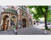 Backsteingebäude mit historischen Architekturdetails wie Rundbögen, verzierten Fenstern und einem Türbogen, teils mit Graffiti bedeckt. Im Vordergrund stehen E-Scooter auf einer gepflasterten Straße, flankiert von Bäumen; rechts ist eine Straßenkreuzung zu sehen.