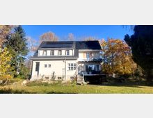 Freistehendes zweigeschossiges Wohnhaus mit hellem Putz, dunklem Mansarddach und mehreren Dachgauben; auf dem Dach sind Solarpaneele montiert. Vor dem Haus liegen ein Garten mit Rasenfläche sowie Bäume mit Herbstlaub rundherum.