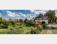 Einfamilienhaus mit großer Glasveranda, umgeben von einem gepflegten Garten mit Bäumen, Blumenbeeten und Rasenflächen; im Vordergrund ein Weg mit gepflastertem Abschnitt und Blumenkästen mit roten Blumen, im Hintergrund eine ländliche Umgebung mit Bäumen.