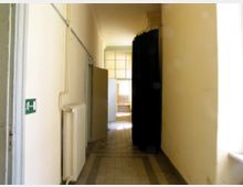 Ein schmaler, gelb-beiger Flur mit hellen Wänden und einem gefliesten Boden; auf der linken Seite befinden sich ein ausgestelltes Notausgangsschild, Heizungsrohre und ein Heizkörper. Am Ende des Flurs steht eine halb geöffnete Tür, durch die Licht in den Raum fällt, daneben hängt ein schwarzer Vorhang.