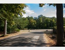 Ein asphaltierter Weg führt durch einen offenen Park mit vereinzelten Bäumen und Sträuchern. Im Hintergrund sind dichte grüne Baumkronen und ein blauer Himmel mit einigen Wolken zu sehen.