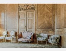 Klassischer Raum mit Holztäfelung an den Wänden, dekorativ geschnitzter Doppeltür und einer Auswahl an gepolsterten Sitzmöbeln, darunter ein rotes Sofa und zwei Sessel mit Blumenmuster auf einem Fischgrät-Holzboden.