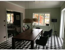 Esszimmer mit Schachbrettmuster-Fliesenboden, einem Holztisch, mehreren gepolsterten Stühlen und einer Hängelampe aus Korbgeflecht; im Hintergrund ein Fenster mit Blick auf einen Garten und ein weiteres Zimmer mit einem Sofa auf der linken Seite.