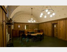 Ein historischer Raum mit Holzvertäfelung, dunklem Boden und einer gewölbten Decke, dekoriert mit dezenten Mustern. Im Vordergrund steht ein hölzerner Tisch mit mehreren Stühlen, im Hintergrund befinden sich ein Tresen und eine Tür neben einem Garderobenständer.