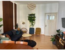 Wohnzimmer mit hellen Wänden und Holzfußboden, ausgestattet mit einer dunklen Eckcouch, zwei bunten Sesseln und einem runden Couchtisch. An der Wand hängen hölzerne Wandpaneele und eine Pflanzeninstallation; eine Tür mit dekorativem Glas und ein Flachbildfernseher sind sichtbar, zusammen mit diversen Zimmerpflanzen und einer goldenen Stehlampe.