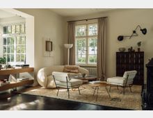 Wohnzimmer mit eleganter Einrichtung: Ein beigefarbenes Sofa, zwei helle Sessel und ein Couchtisch stehen auf einem gemusterten Teppich. Große Sprossenfenster mit beigefarbenen Vorhängen lassen Tageslicht herein, während ein dunkler Holzboden und eine dekorative Kommode den Raum abrunden.