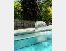 Ein Außenpool mit klarem, türkisfarbenem Wasser und einem kleinen Wasserfall aus einem Edelstahlbogen. Im Hintergrund sind eine Steinmauer und dichte, grüne Bepflanzung zu sehen.