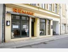 Außenansicht eines gelb gestrichenen Gebäudes mit großen Glasfenstern und einem Eingang in der Mitte. Über den Fenstern und der Tür ist ein Schild mit der Aufschrift „REMAKE Wein, Genuss und Emotion“ angebracht.