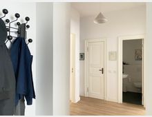 Heller Wohnungsflur mit weiß gestrichenen Wänden, Holzboden und einer Deckenleuchte, mit mehreren weißen Zimmertüren. Links hängt eine Garderobe an einer schwarzen Wandhakenleiste, rechts ist eine geöffnete Tür zu einem kleinen Bad mit Waschbecken und Toilette zu sehen.