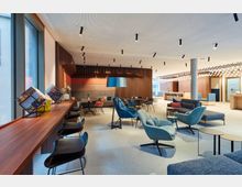 Moderne Lounge mit einer offenen, einladenden Einrichtung, bestehend aus bequemen Sesseln, Sofas und kleinen Tischen. Die Atmosphäre wird durch warme Holzwände, farbige Polster, große Fenster und dezente Deckenbeleuchtung ergänzt.